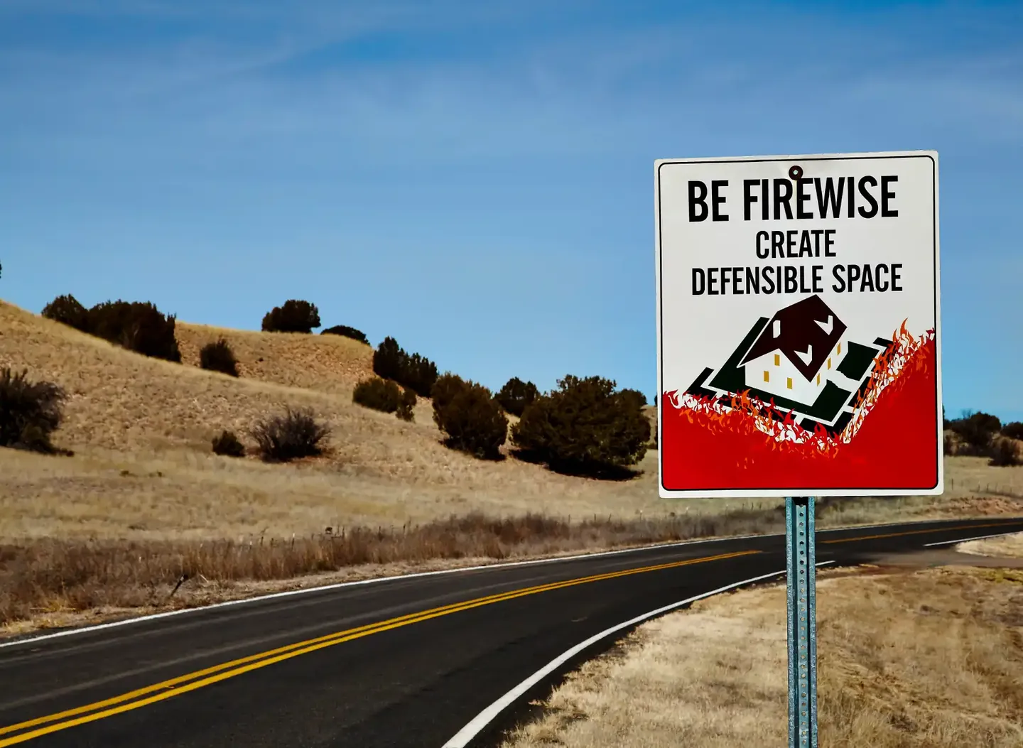 Roadside sign encouraging defensible space near grass-covered hills