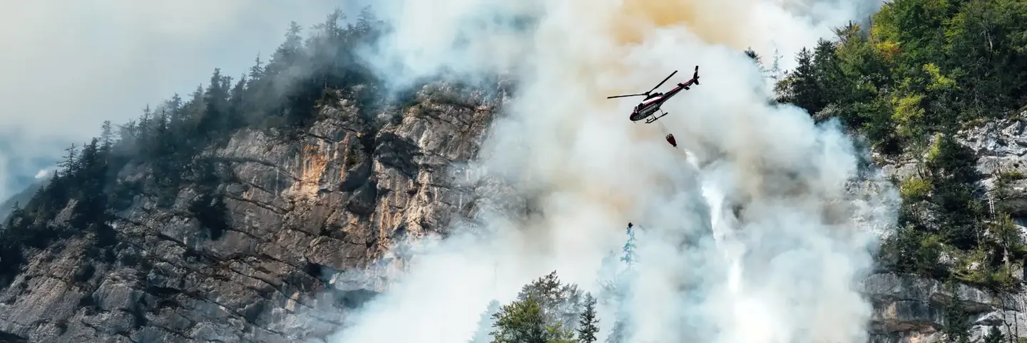 Helicopter carrying a water bucket above a smoky wooded hillside